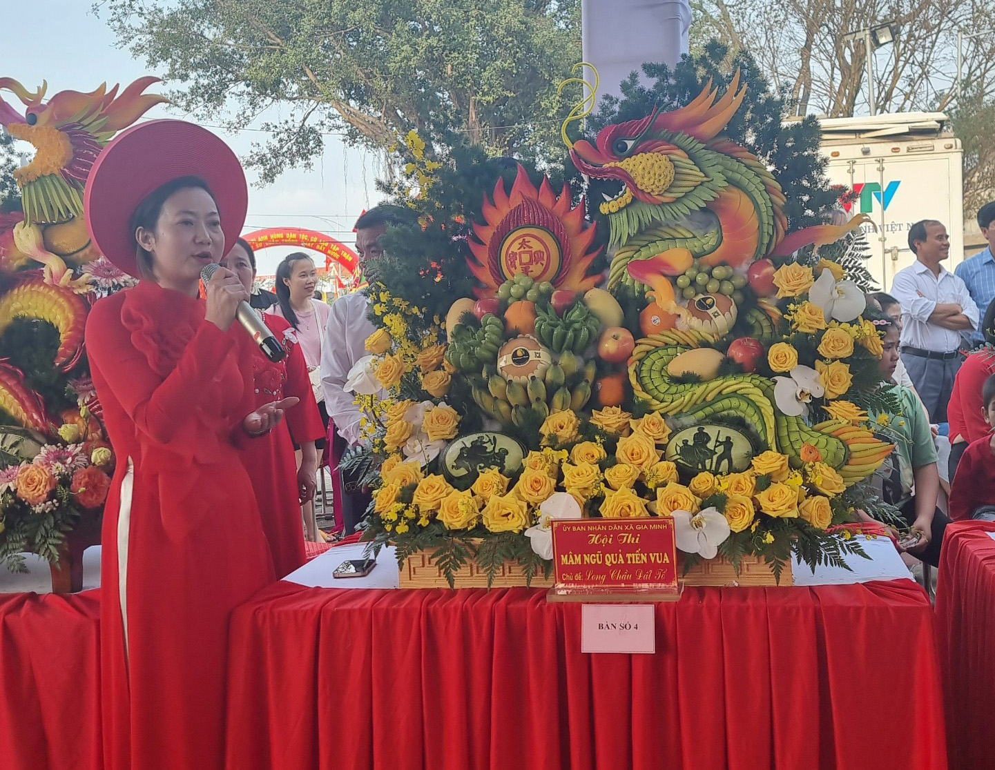 A person in red dress and hat standing next to a table with flowers Description automatically generated